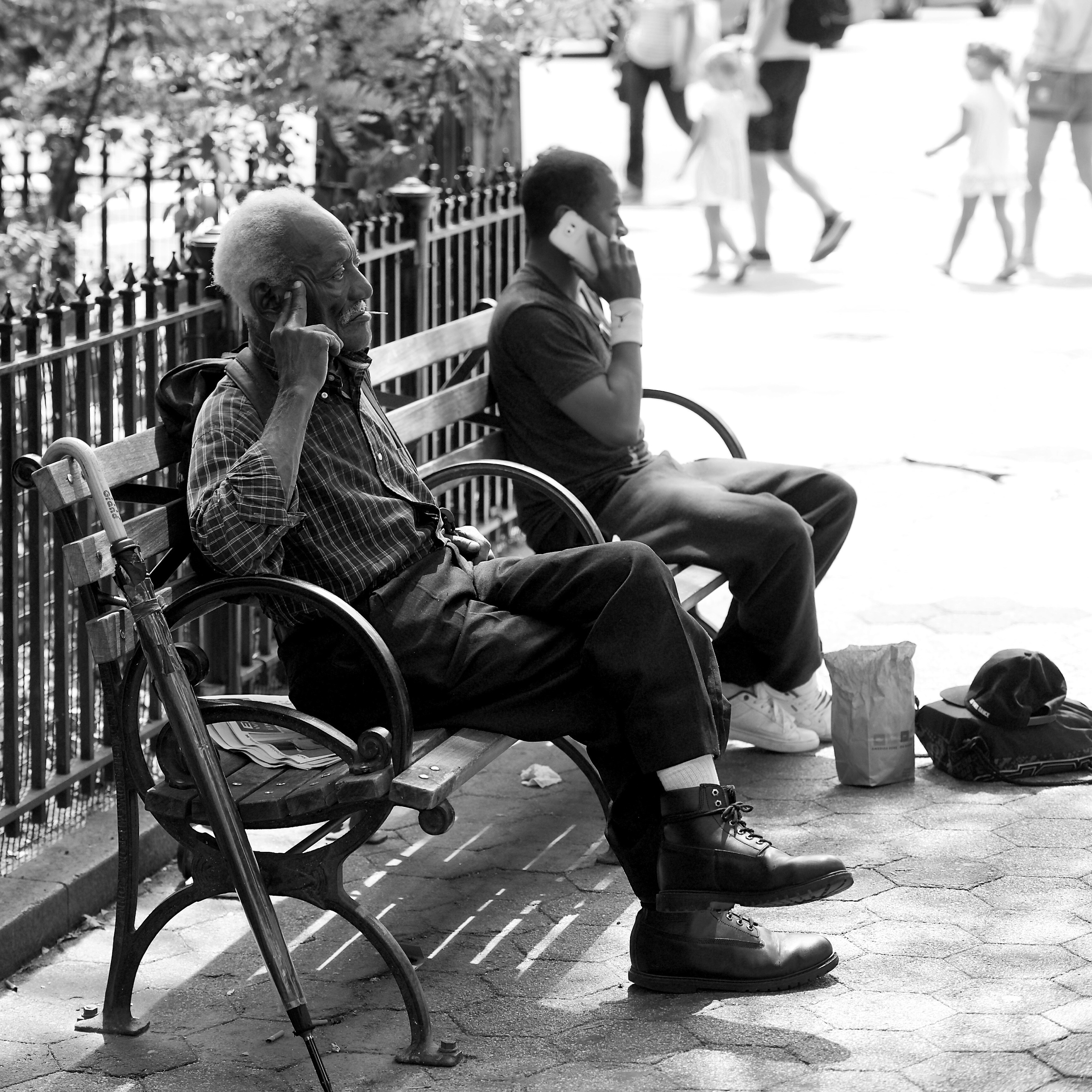 Two men on a bench mirror each other’s private calls in a blown-bright city park.