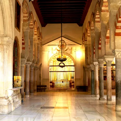 A hanging lamp between rows of columns and red-and-white arches, facing a brightly lit barred doorway reflected on the stone floor.