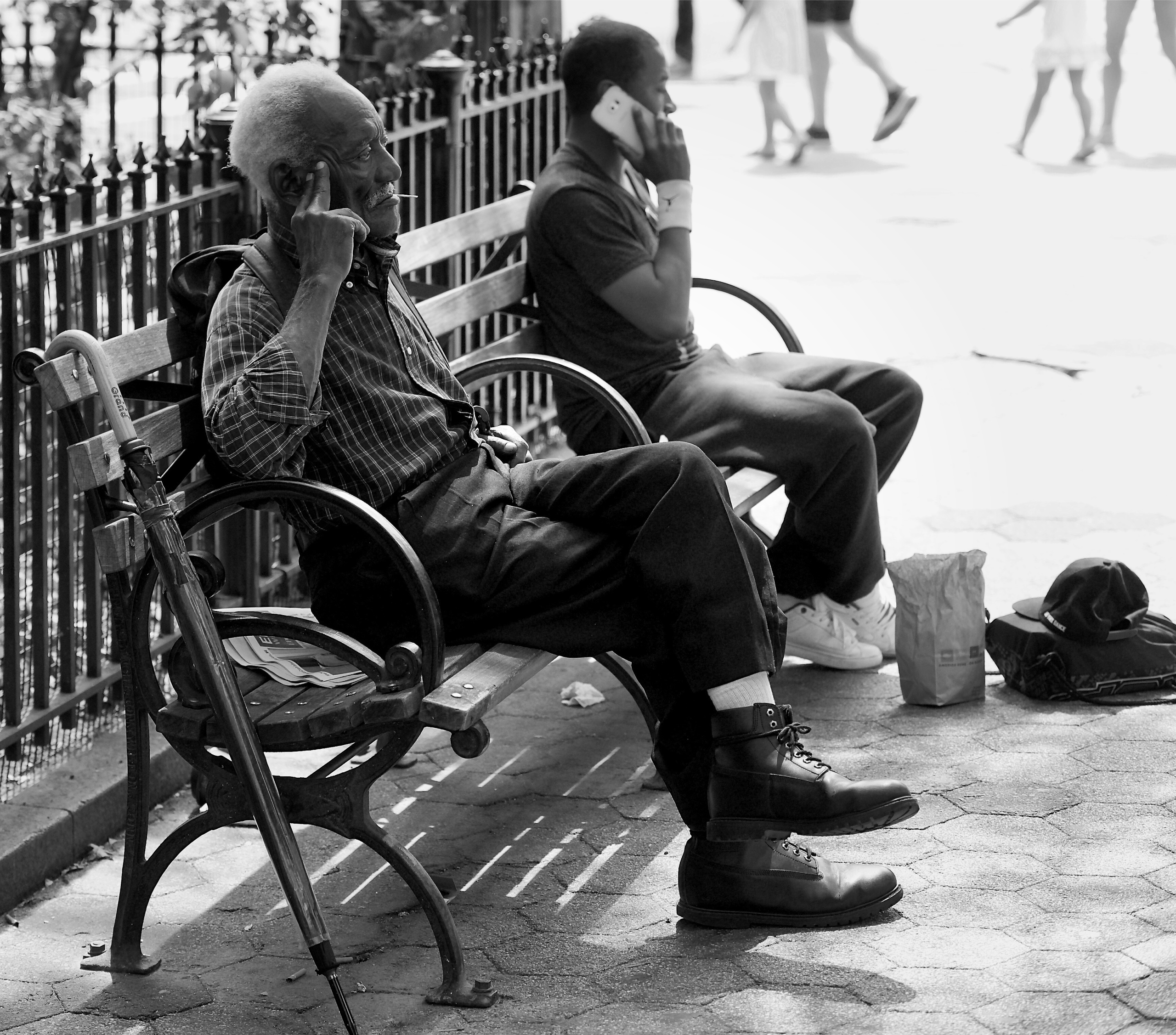Edited preview of Two men on a bench mirror each other’s private calls in a blown-bright city park.